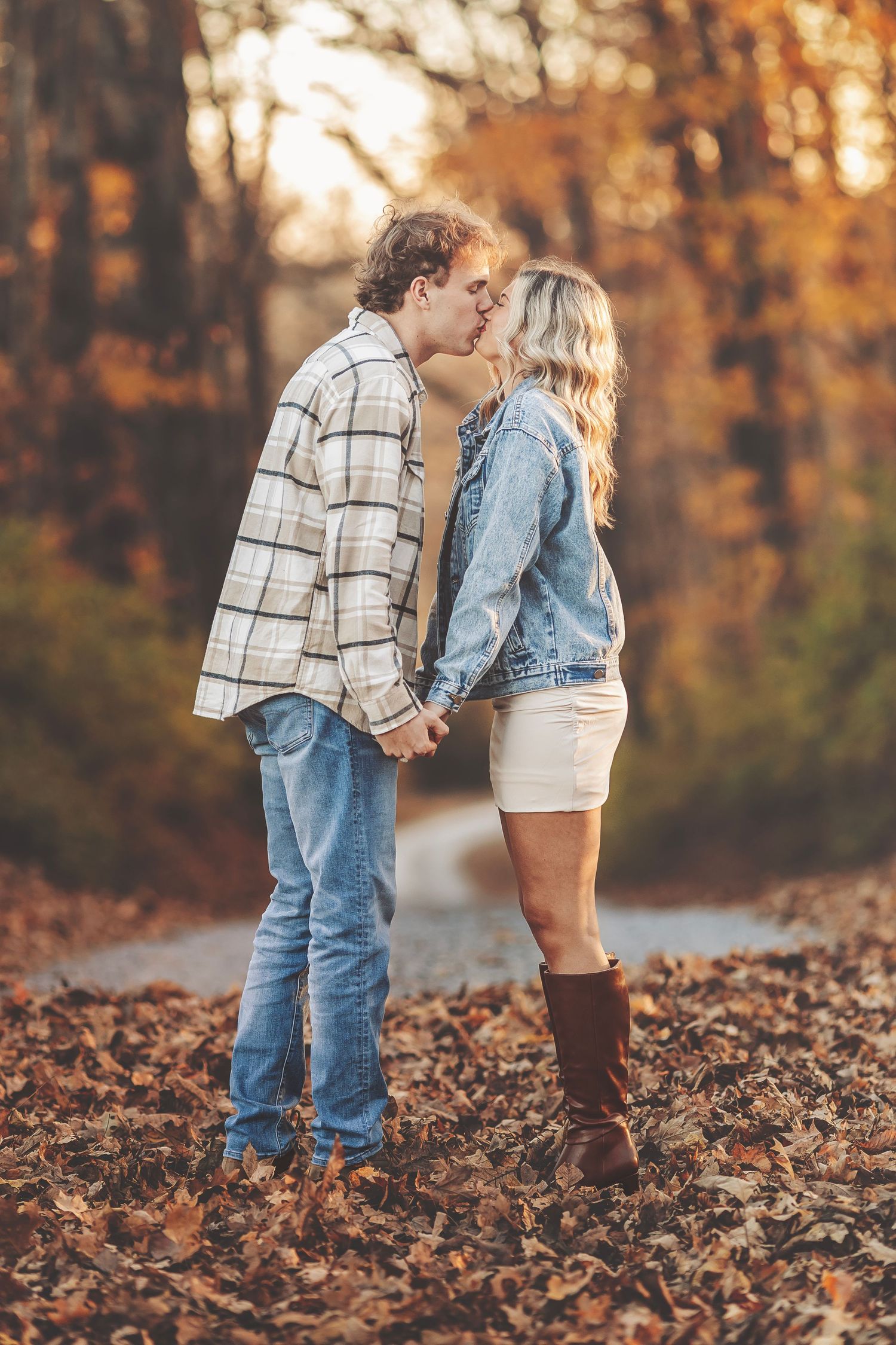 Couple embracing and smiling in a sunlit autumn forest, golden light filtering through bare trees onto fallen leaves.