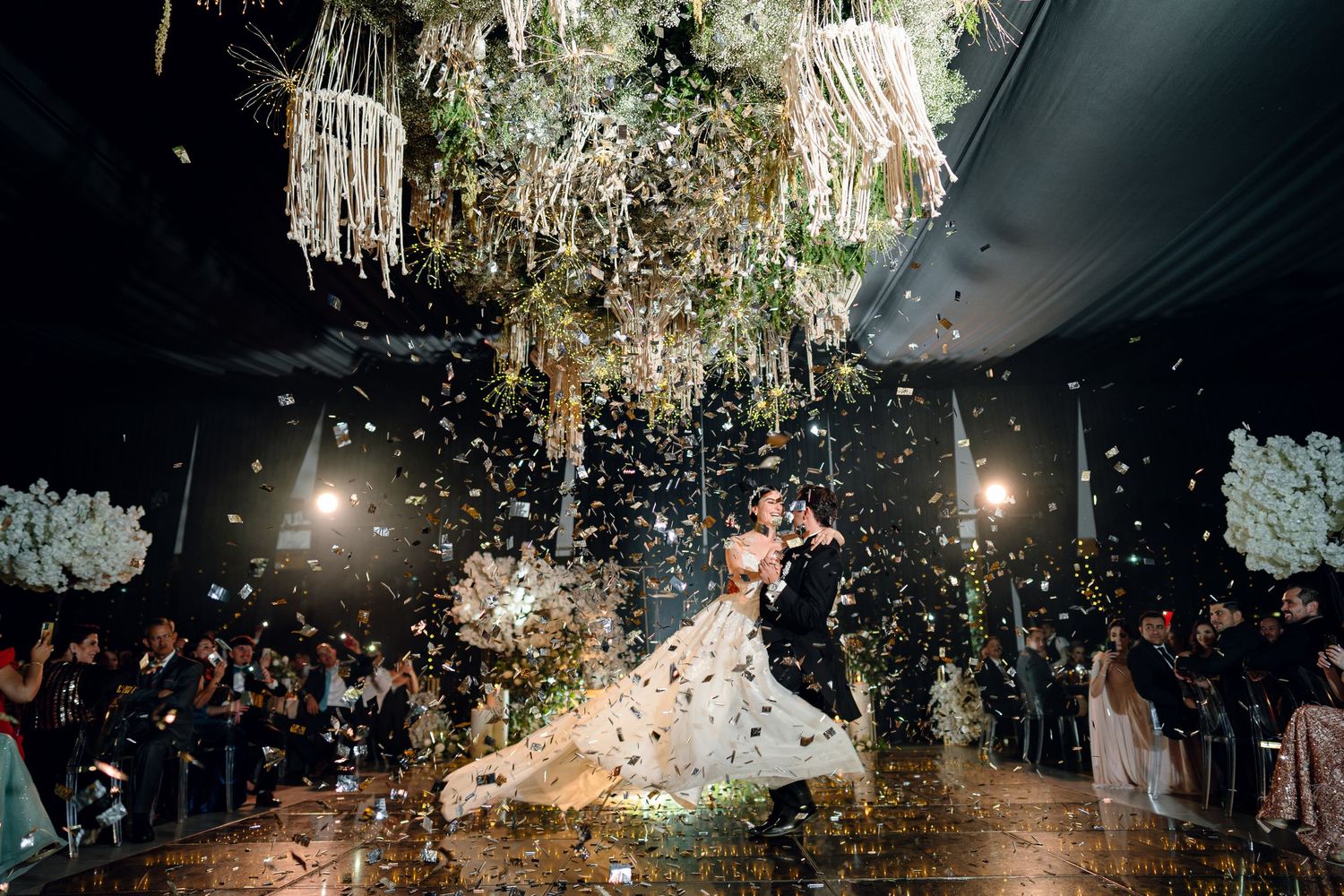 Dynamic wedding celebration photo with falling confetti and hanging floral decorations in dark reception venue.