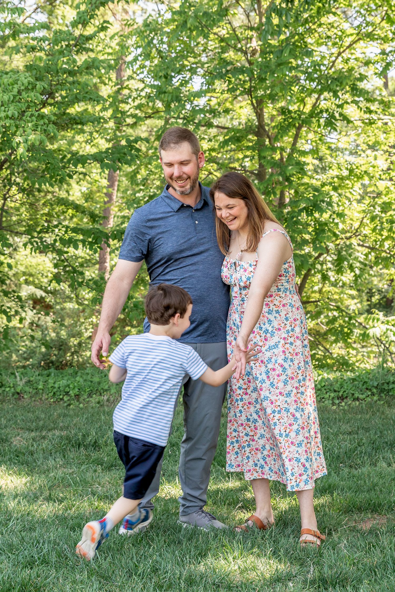 A happy family of three enjoys a sunny day outdoors, with parents smiling as their young son runs toward them on green grass.