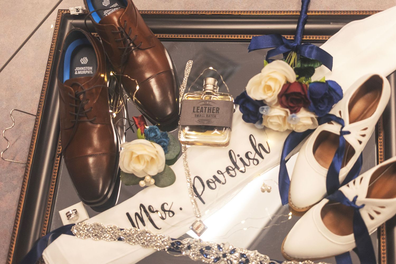 Wedding flat lay with brown dress shoes, white bridal heels, blue roses, perfume, and a Mrs. Povolish sash on a mirror tray.