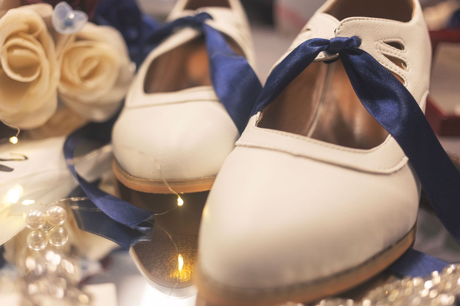 White bridal flats with navy blue ribbon bows beside cream roses and glowing candles for a wedding.
