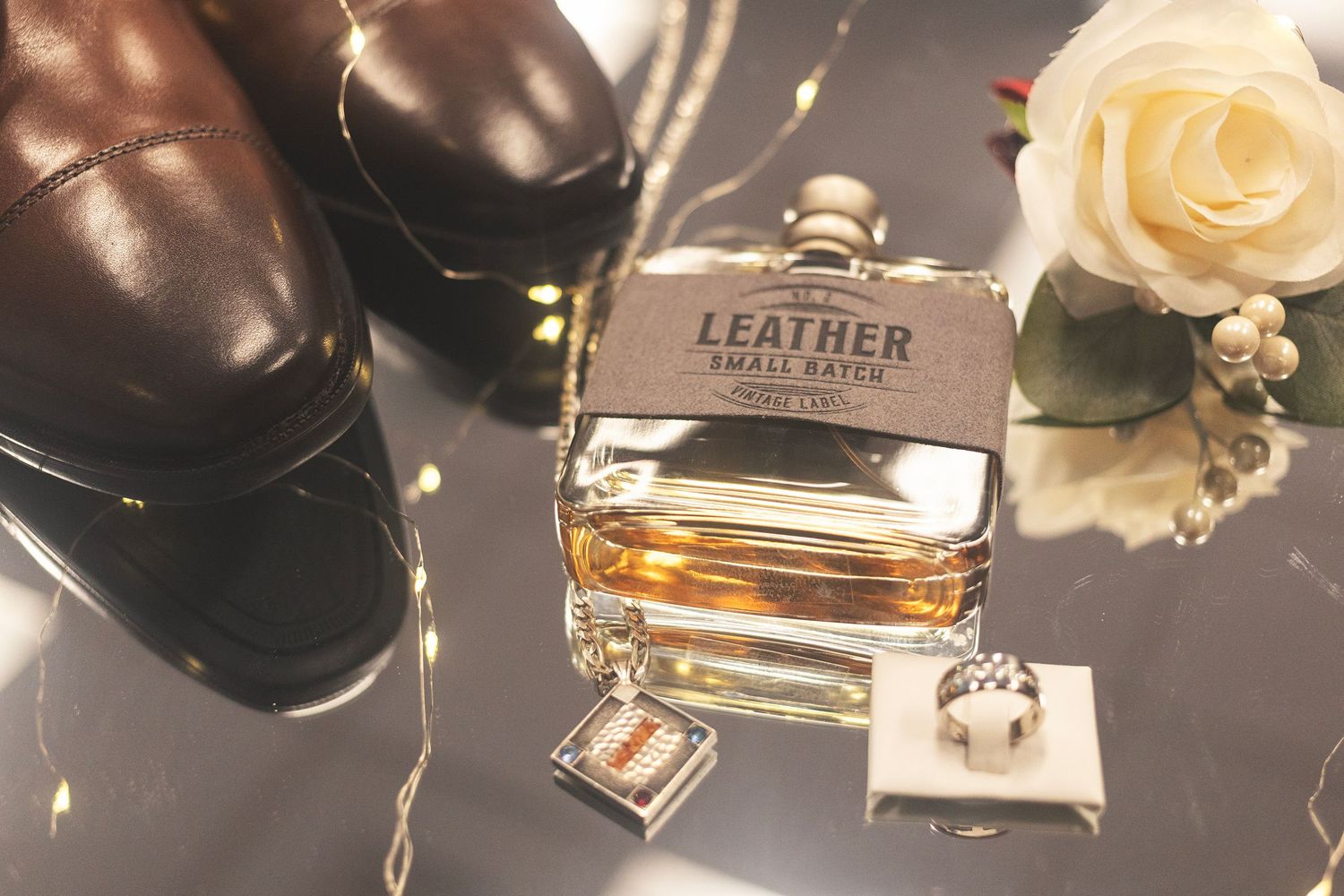 Men's wedding flat lay with leather cologne, brown dress shoes, cufflinks, and a ring on a reflective surface.