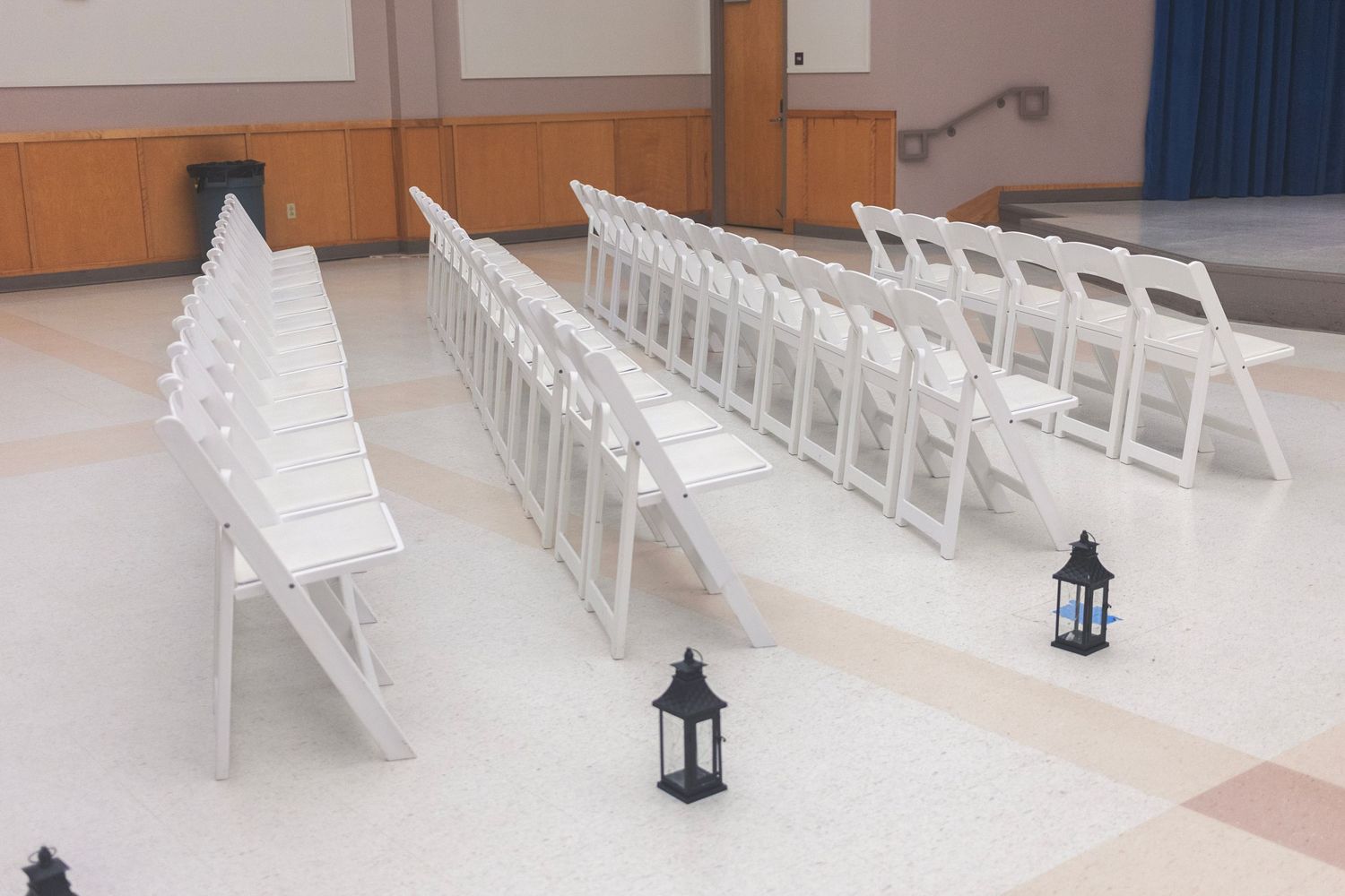 Rows of white folding chairs with black lanterns on floor, set up for an elegant indoor ceremony or event.