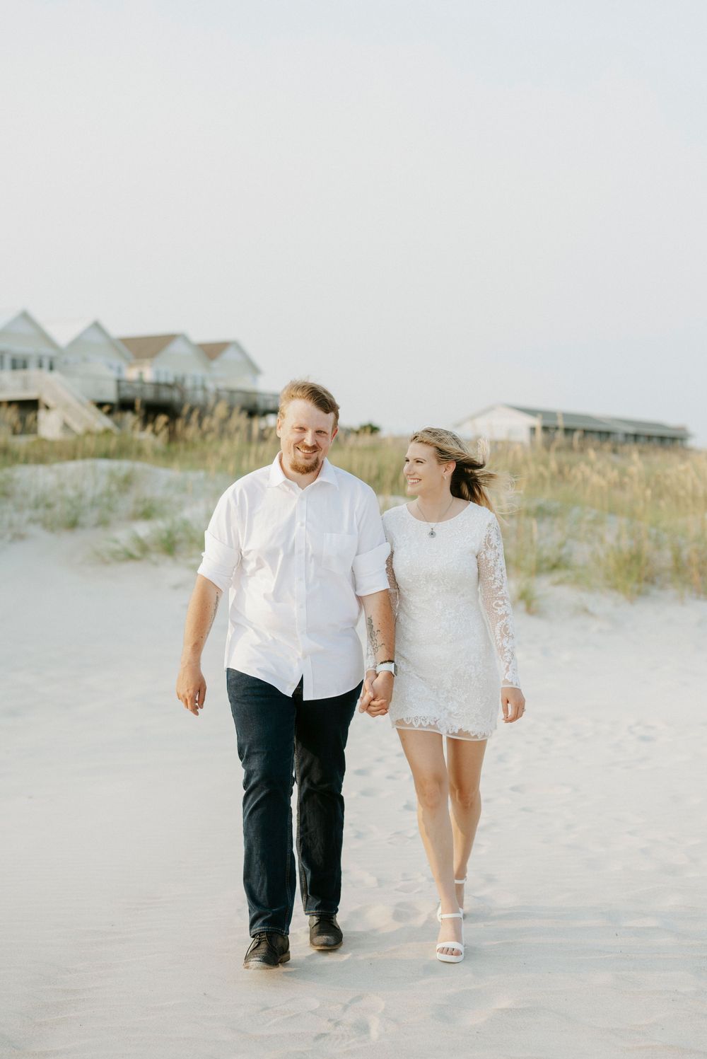 Couple in white outfits walking hand in hand on a sandy beach with beach houses in the background.