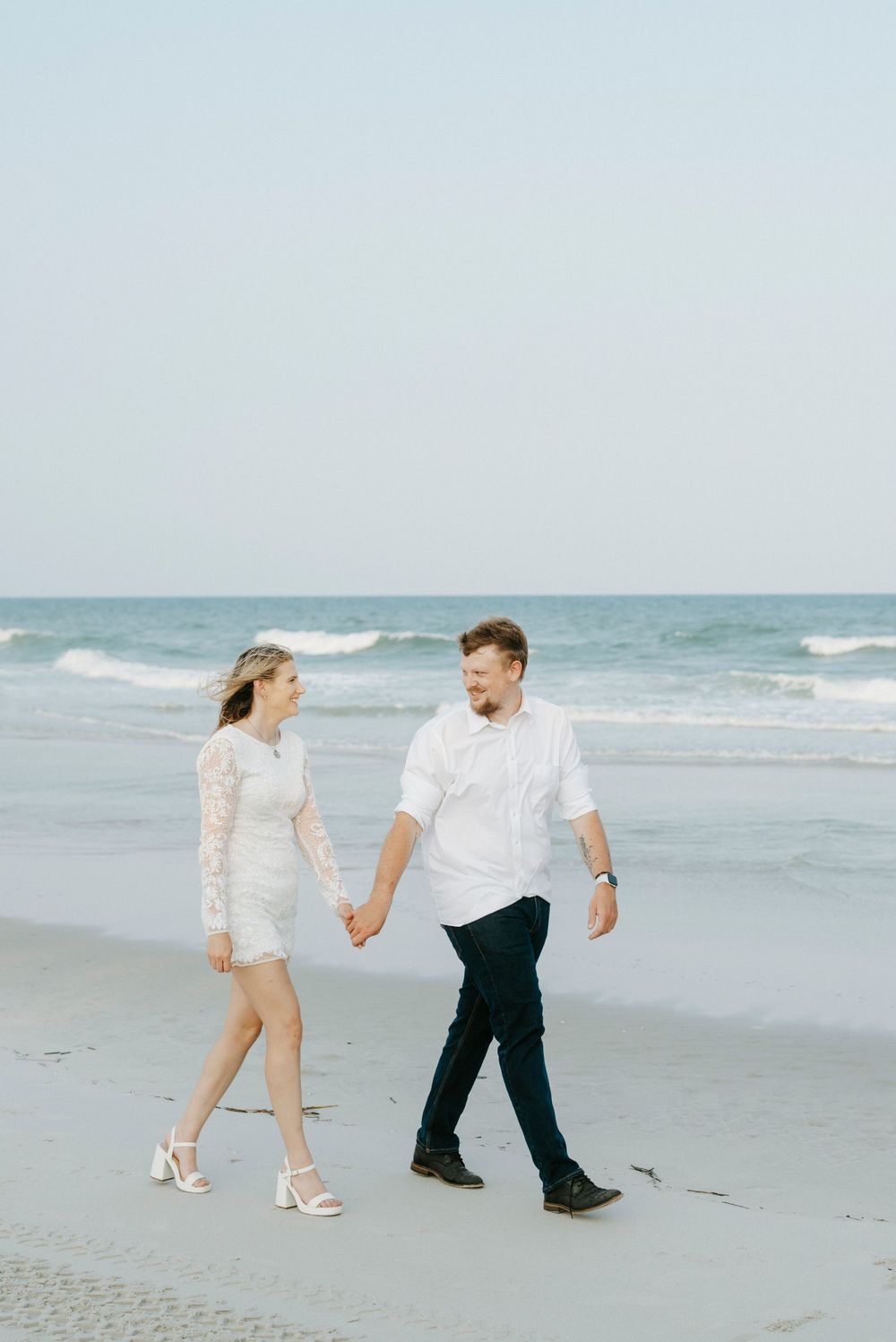 Couple holding hands walking on beach, woman in white lace dress and man in white shirt, ocean waves behind them.