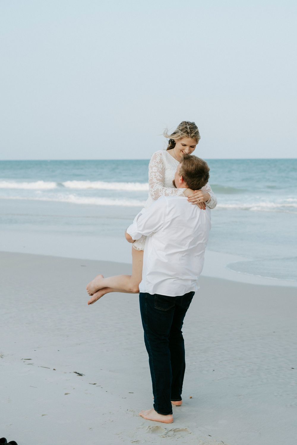 A groom lifts his bride on a sandy beach, both dressed in white, with ocean waves in the background.