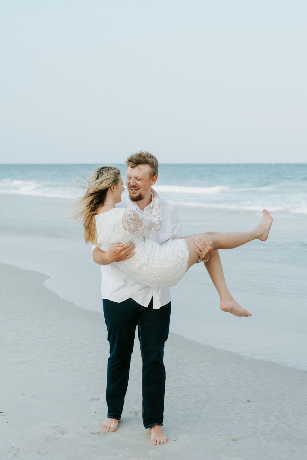 A man carries a laughing woman in his arms on a sandy beach, both wearing white, with ocean waves behind them.