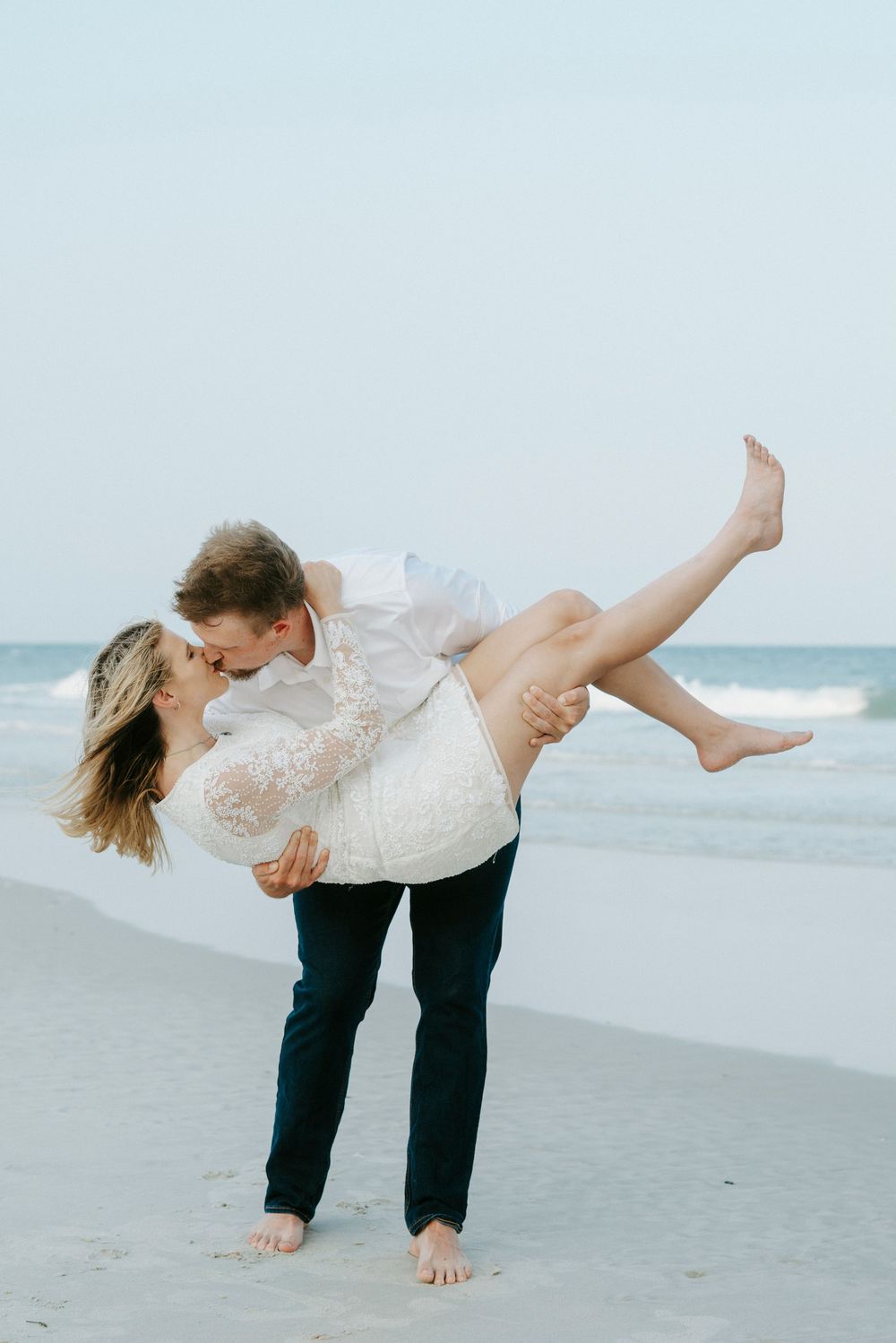 A couple shares a romantic kiss on the beach, as the man lifts the woman in a white lace dress by the ocean.