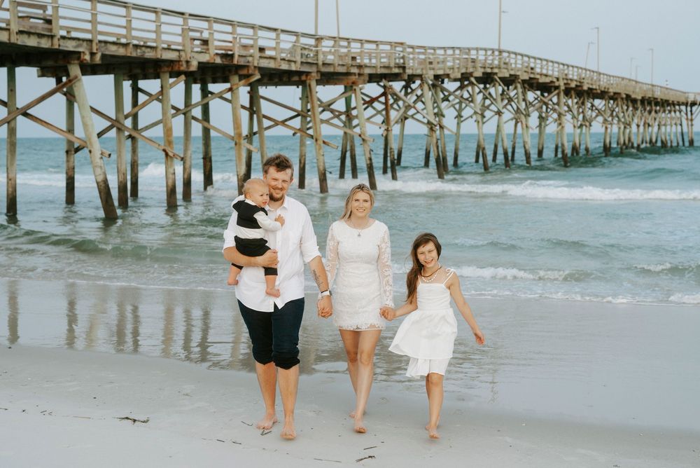 Family of four walking on beach near wooden pier, parents holding hands with children in white outfits on sandy shore.