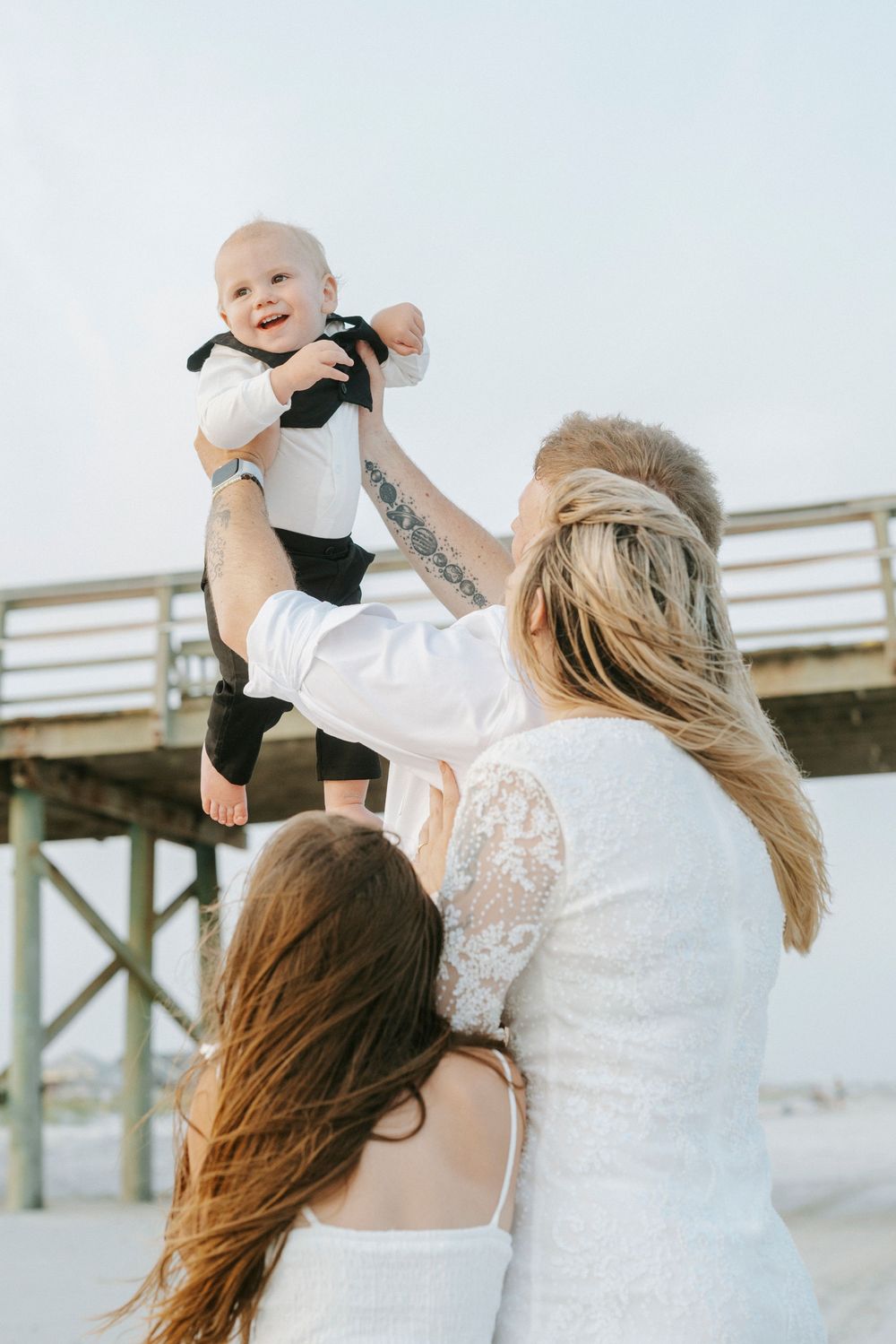 Parents lift a smiling baby in black and white outfit above their heads near a beach pier on a sunny day.