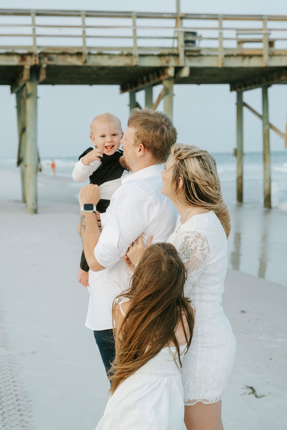 Family with baby and young girl posing happily on beach near wooden pier, parents dressed in white.