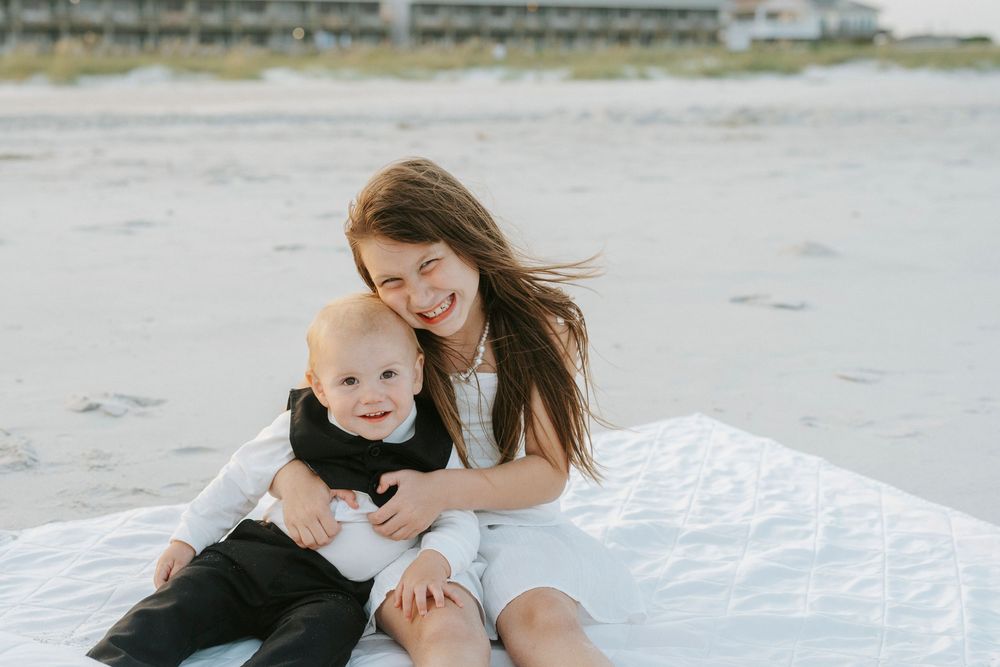 A smiling mother in a white dress sits on a beach blanket with her baby boy in a black outfit.