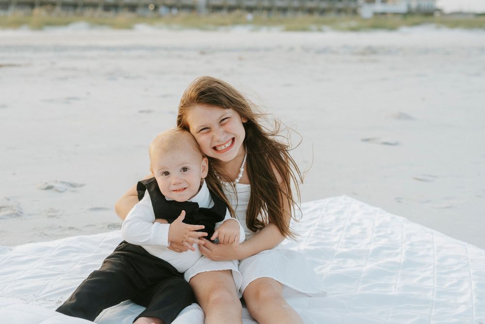 A smiling girl in white dress hugs a baby boy in black suit on a white blanket at the beach.