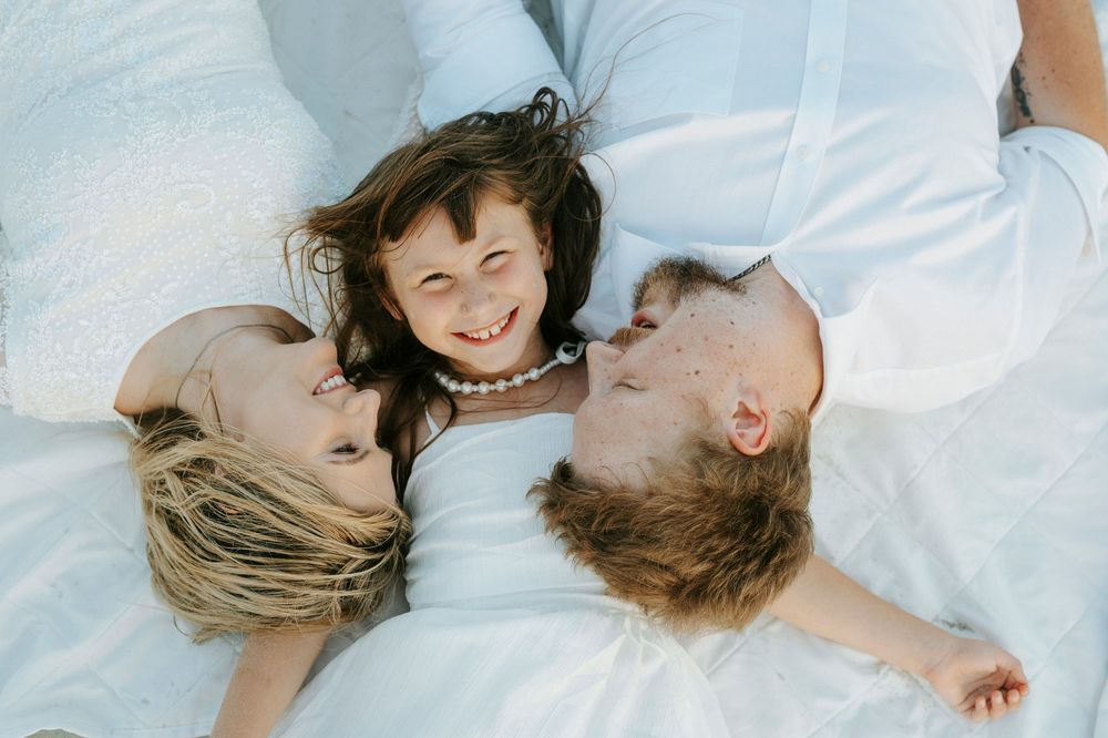 A joyful family of three lying on white fabric, parents kissing their smiling young daughter from both sides.