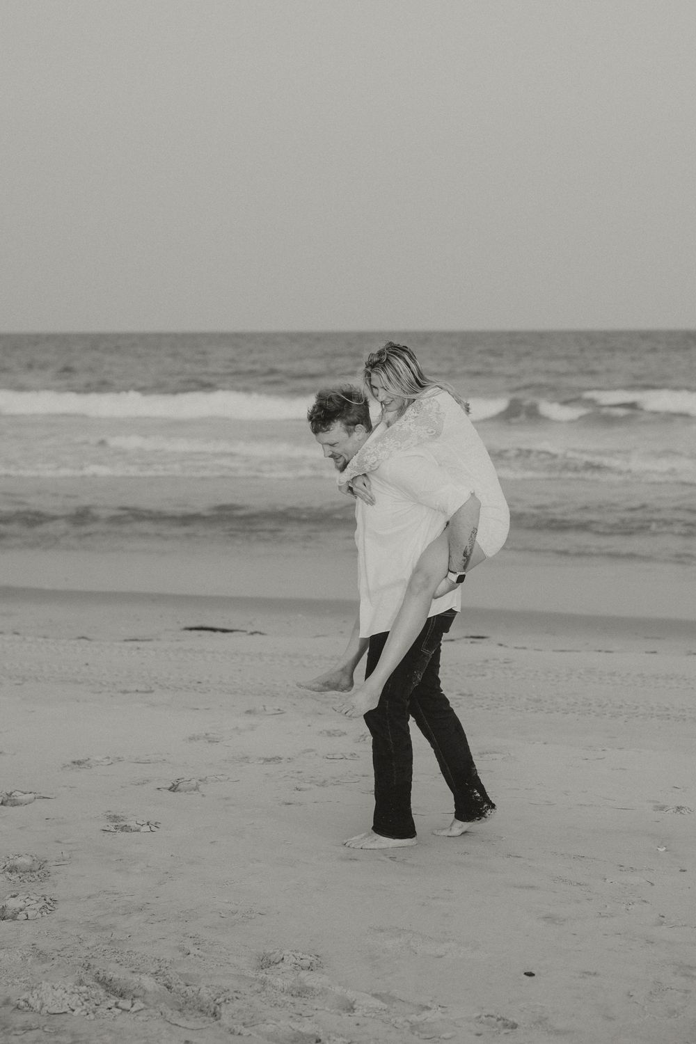 Black and white photo of a couple on the beach, man giving woman a piggyback ride by the ocean waves.