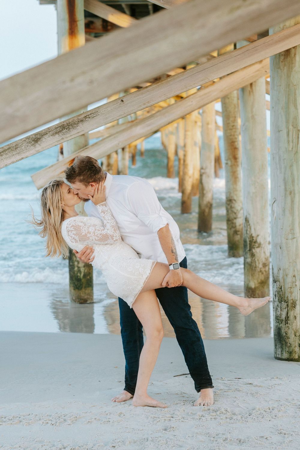 Couple sharing a romantic kiss under a beach pier, man dipping woman in white lace dress on sandy shore.