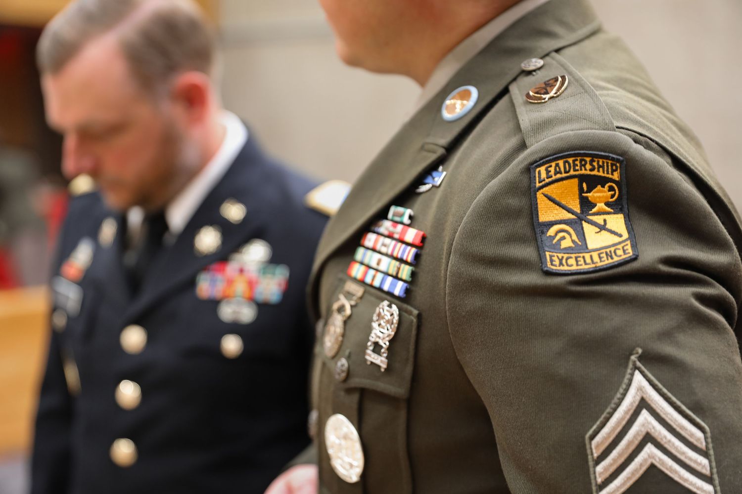 Two military officers in uniform, one displaying Leadership Excellence patch and service ribbons on olive green Army dress uniform.