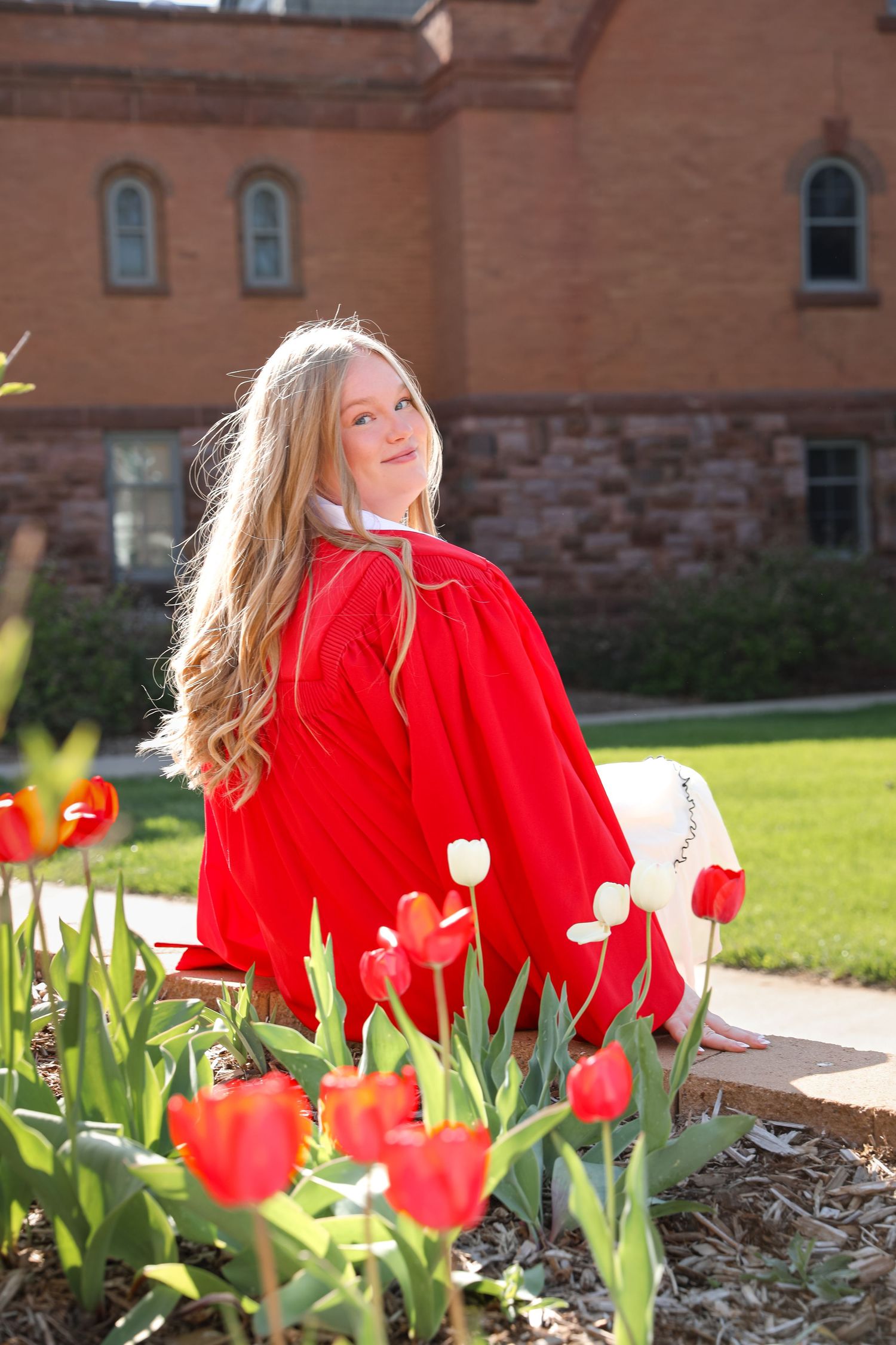 Young girl in red graduation gown sits among red tulips outside a brick building on a sunny day.