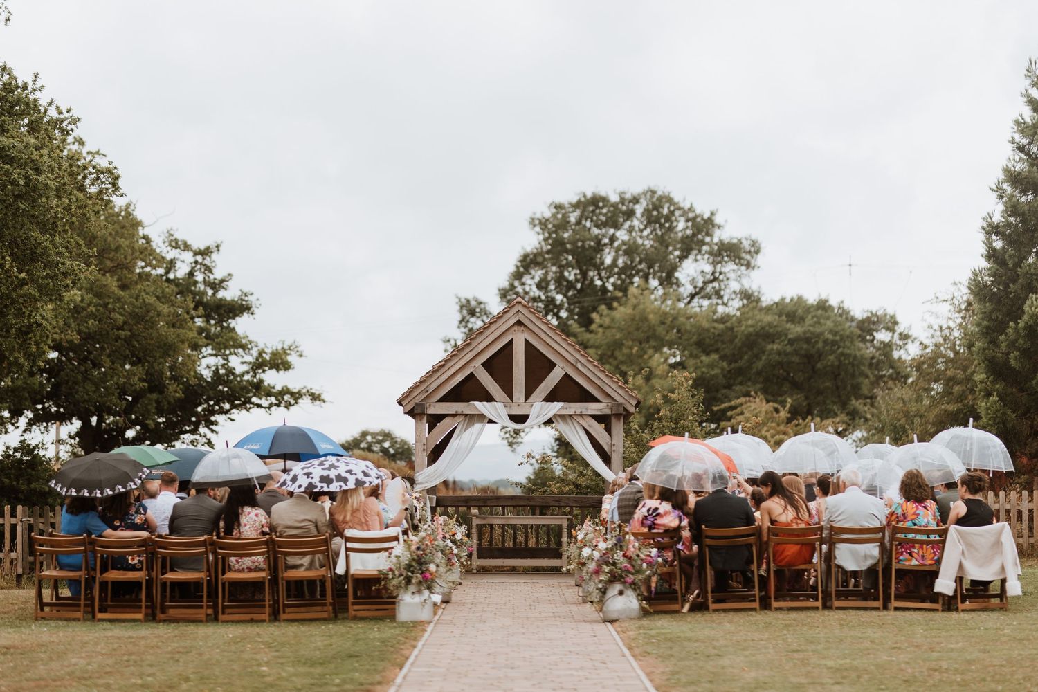 Outdoor wedding ceremony at Moat Hall Barwith guests holding umbrellas seated before a rustic wooden arch in a lush garden setting.