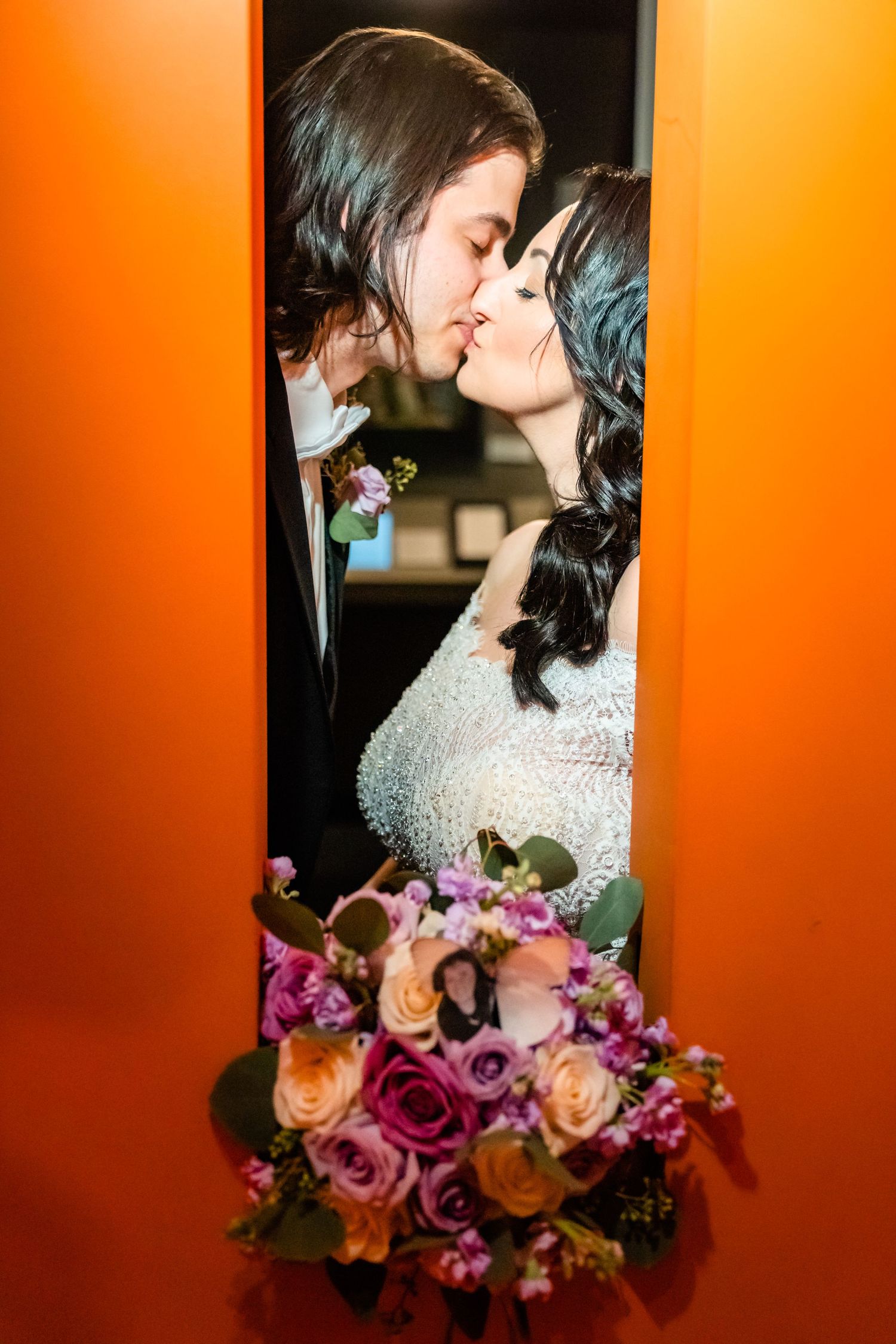 A bride and groom share a romantic kiss framed by orange walls, bride holding a colorful pink and peach rose bouquet.