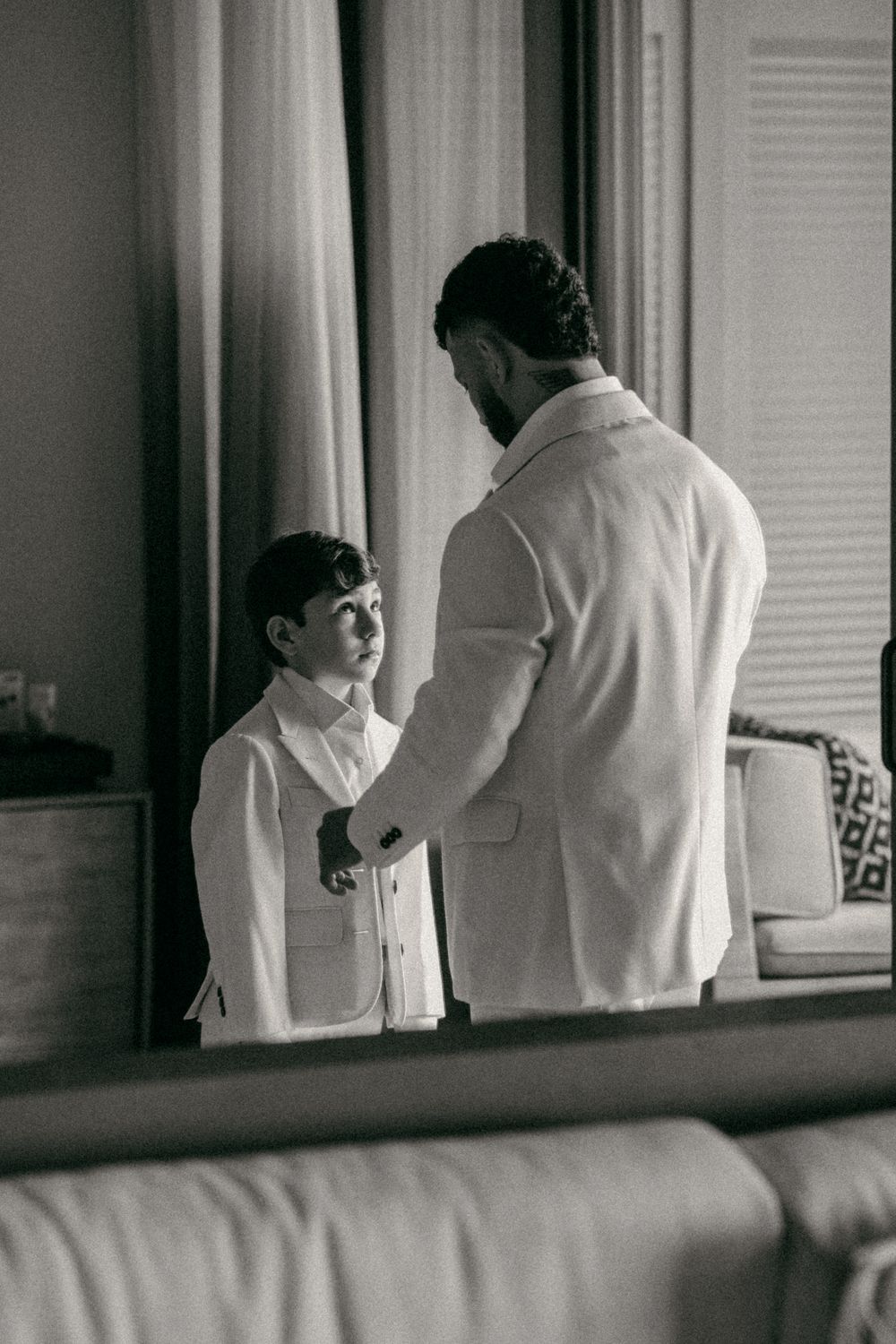 Black and white photo of a man in a suit jacket adjusting a young boy's formal outfit indoors.