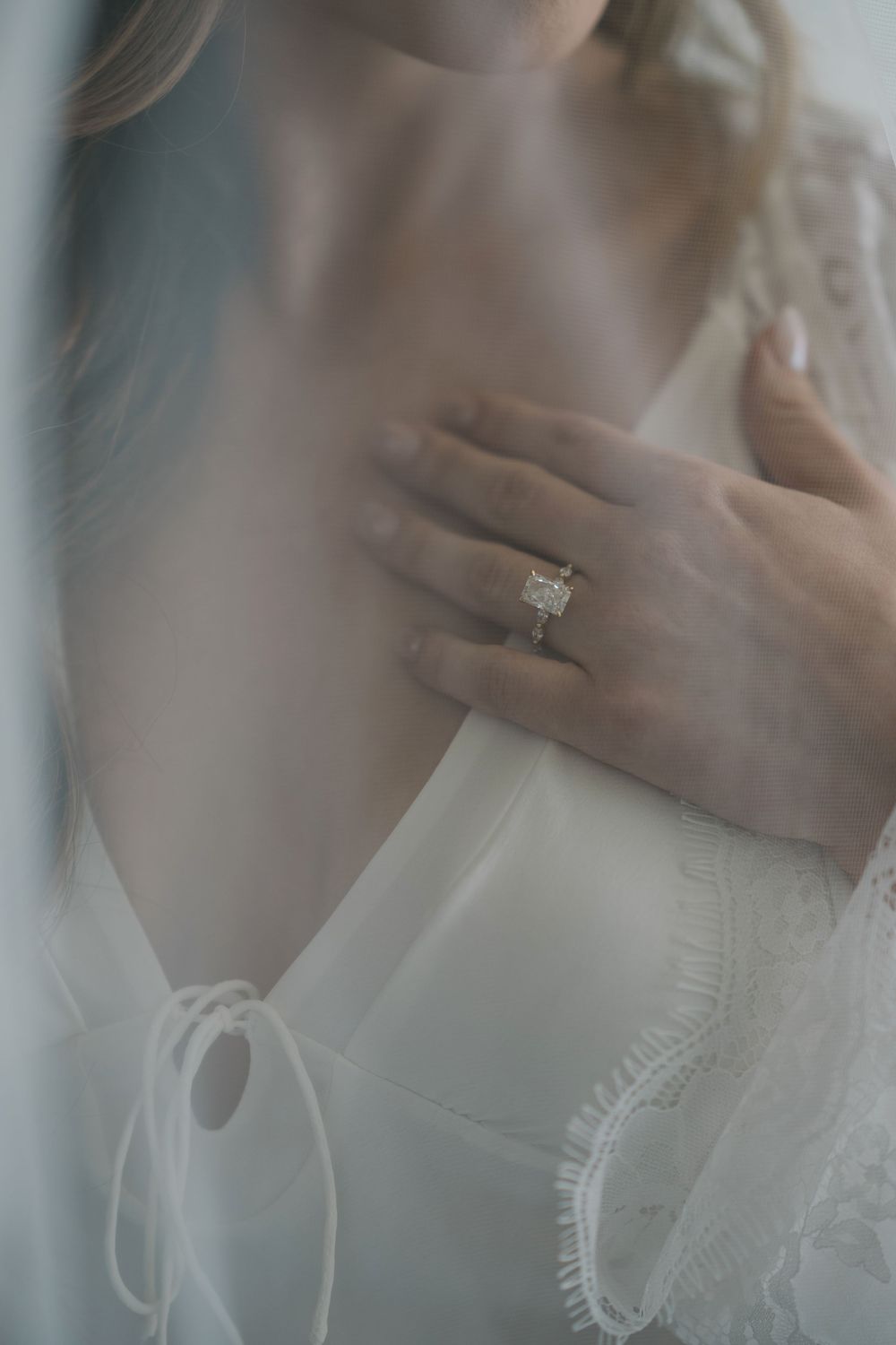 Bride in white lace robe displaying elegant diamond engagement ring on her hand against her chest.
