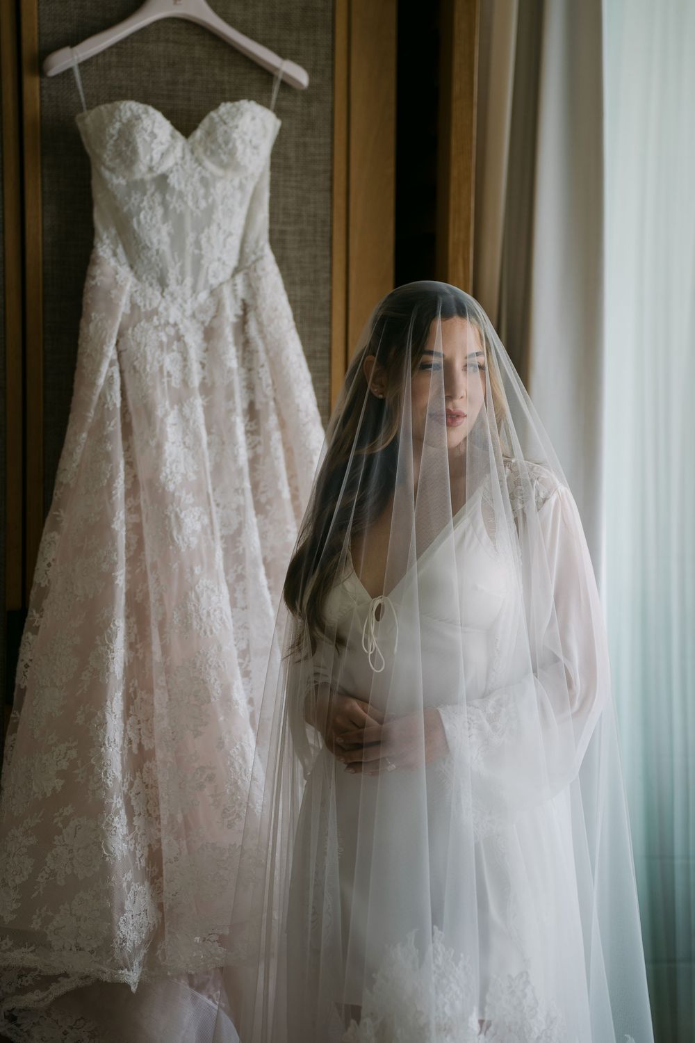 Bride in white robe and veil stands beside hanging lace wedding dress near window in bridal suite.