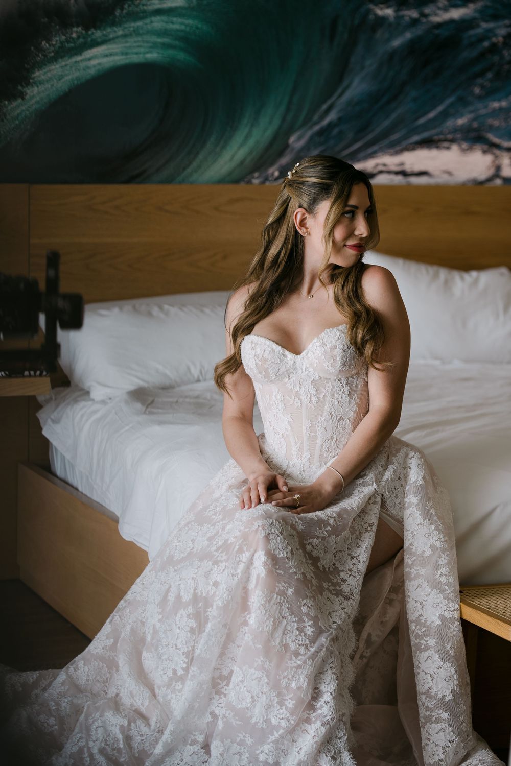 A smiling bride in a strapless lace wedding gown sits on a hotel bed with ocean wave artwork behind her.