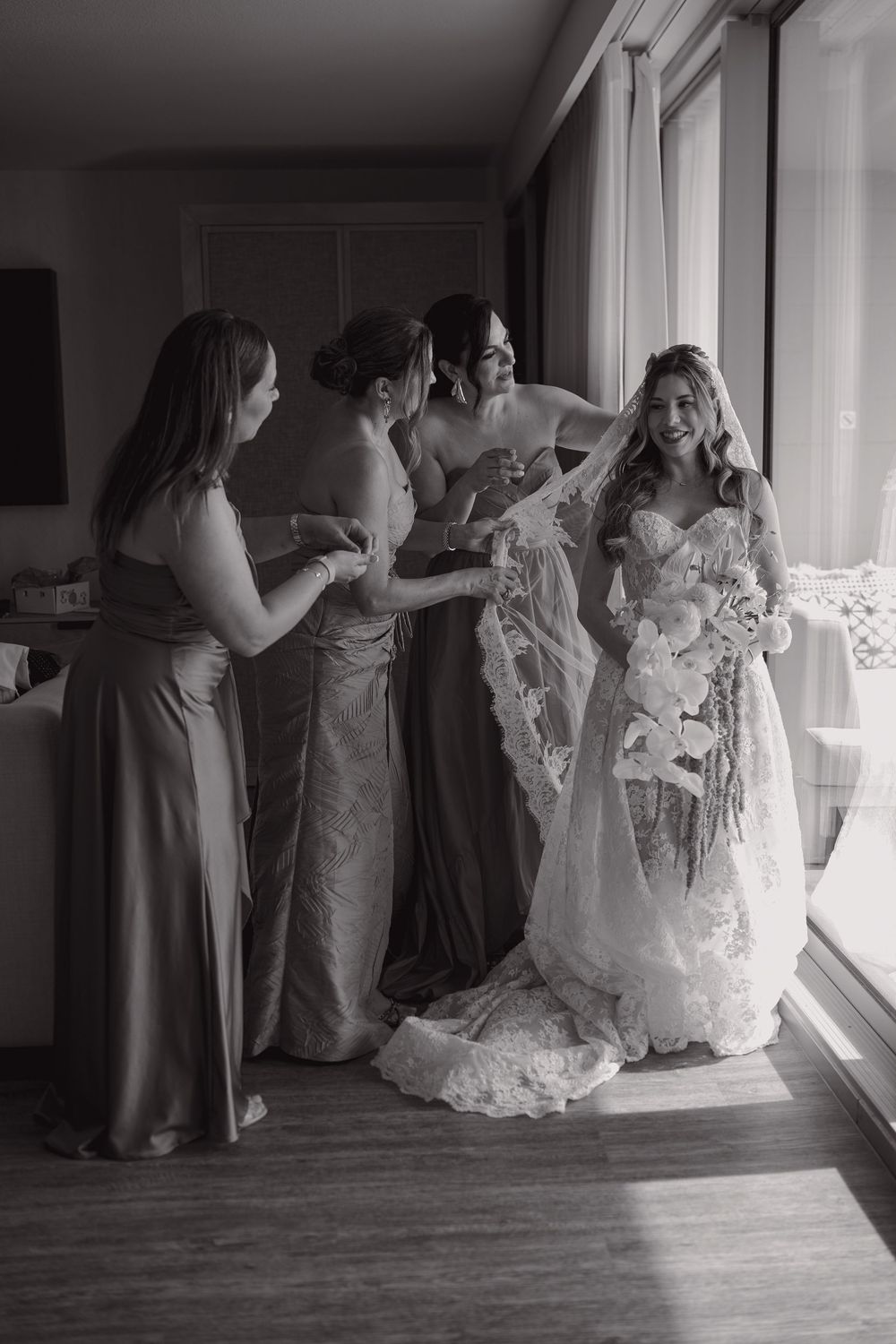 Bride in lace gown holds bouquet as bridesmaids adjust veil near window in black and white photo.