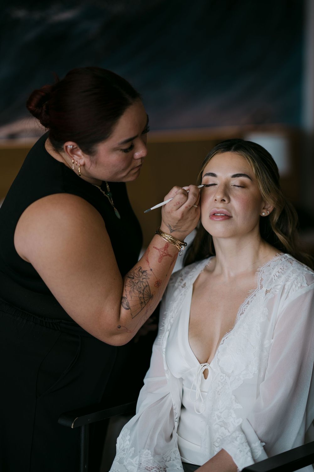 Makeup artist applying eyeshadow to a bride in a white lace robe during wedding preparations.