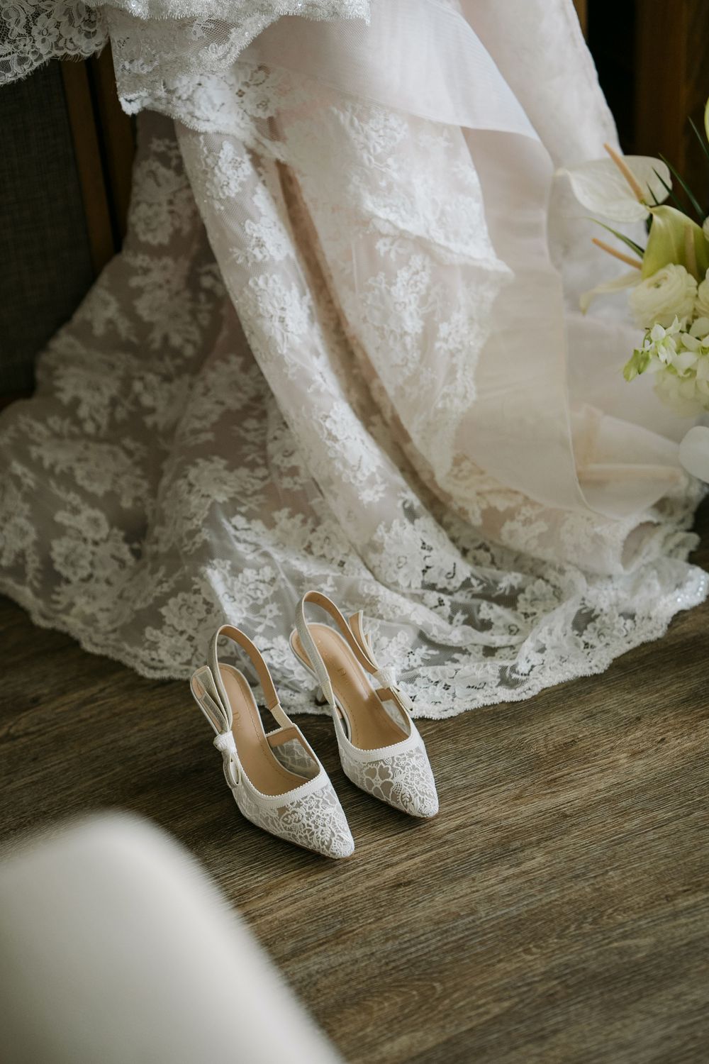 White lace bridal heels on wooden floor beside an elegant lace wedding dress and floral bouquet.