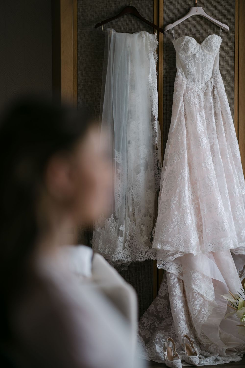 Bride gazing at two elegant lace wedding dresses hanging on display, one blush pink and one white, in a softly lit room.