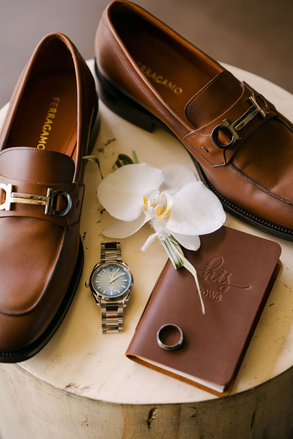 Ferragamo brown leather loafers with gold hardware, watch, wedding ring, leather journal, and white orchid on wooden surface.