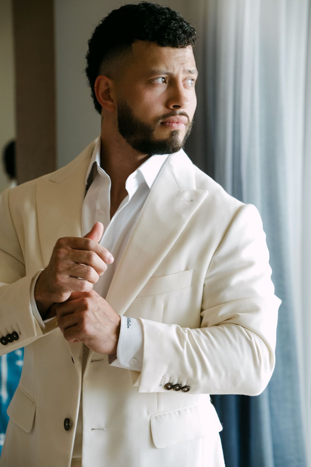 A well-dressed man in a cream white suit adjusts his cufflinks while gazing thoughtfully to the side.