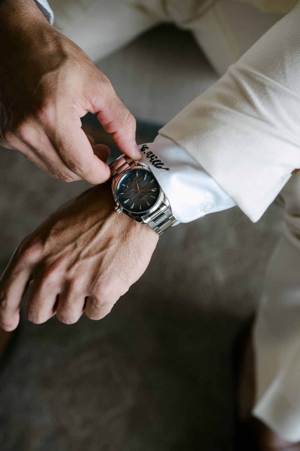 Man adjusting a silver wristwatch on his wrist while wearing a white dress shirt with script embroidery on the cuff.