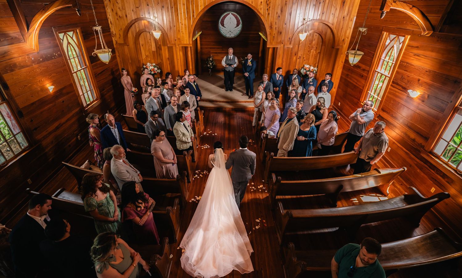 Aerial view of a bride in a white gown walking down the aisle of a wooden chapel filled with wedding guests.