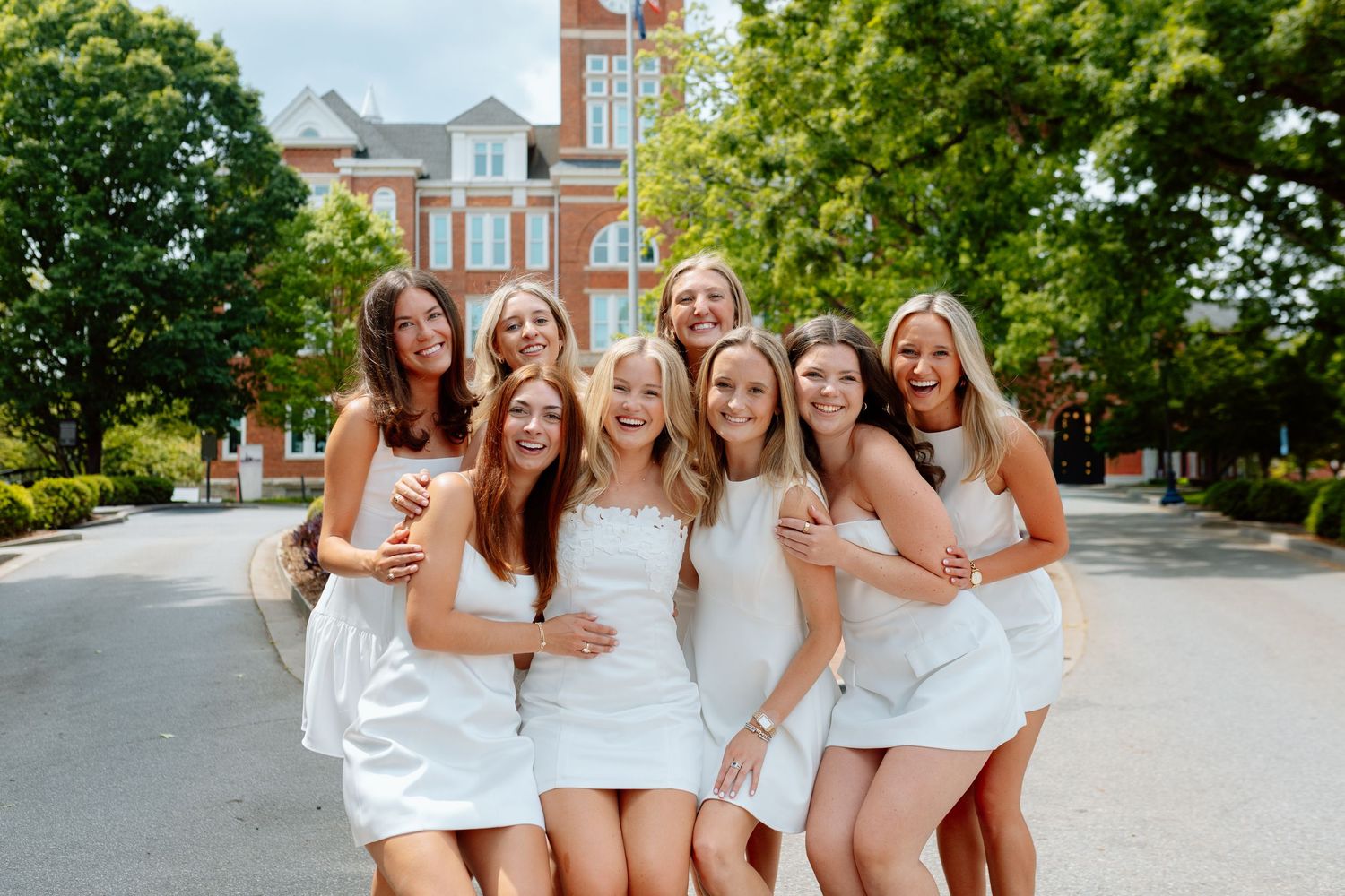 Group of young women in matching white dresses smiling and posing together outdoors on a sunny campus.