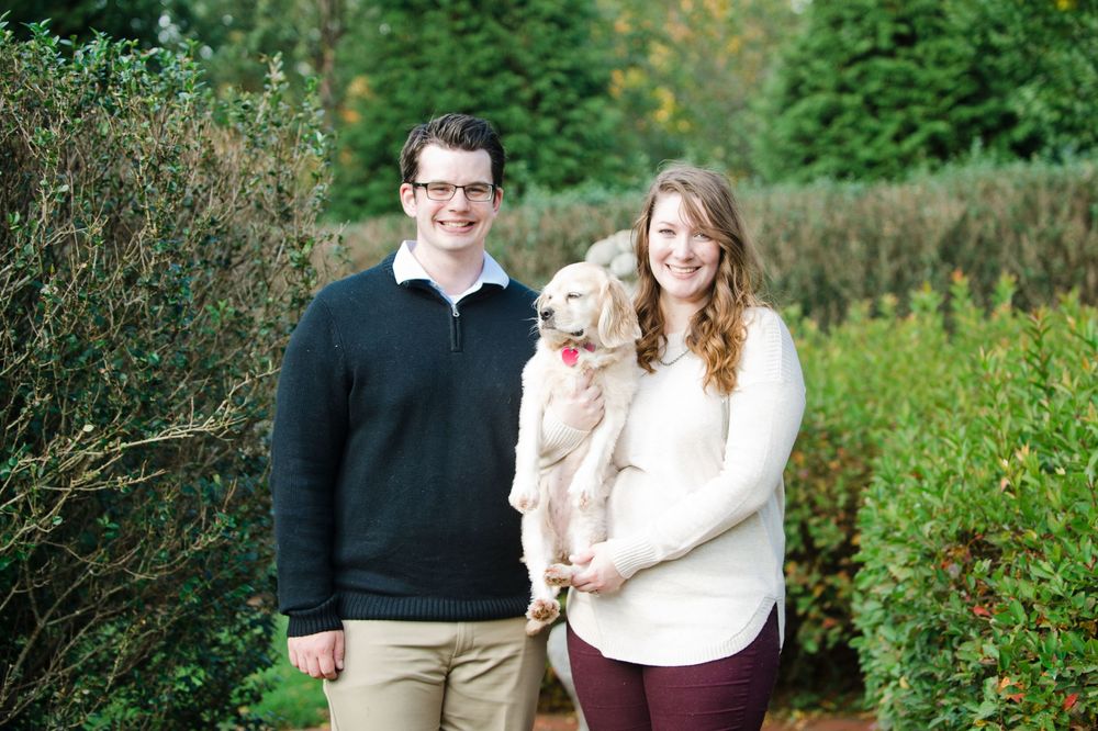 Smiling couple standing together holding a golden puppy in a lush green garden setting.