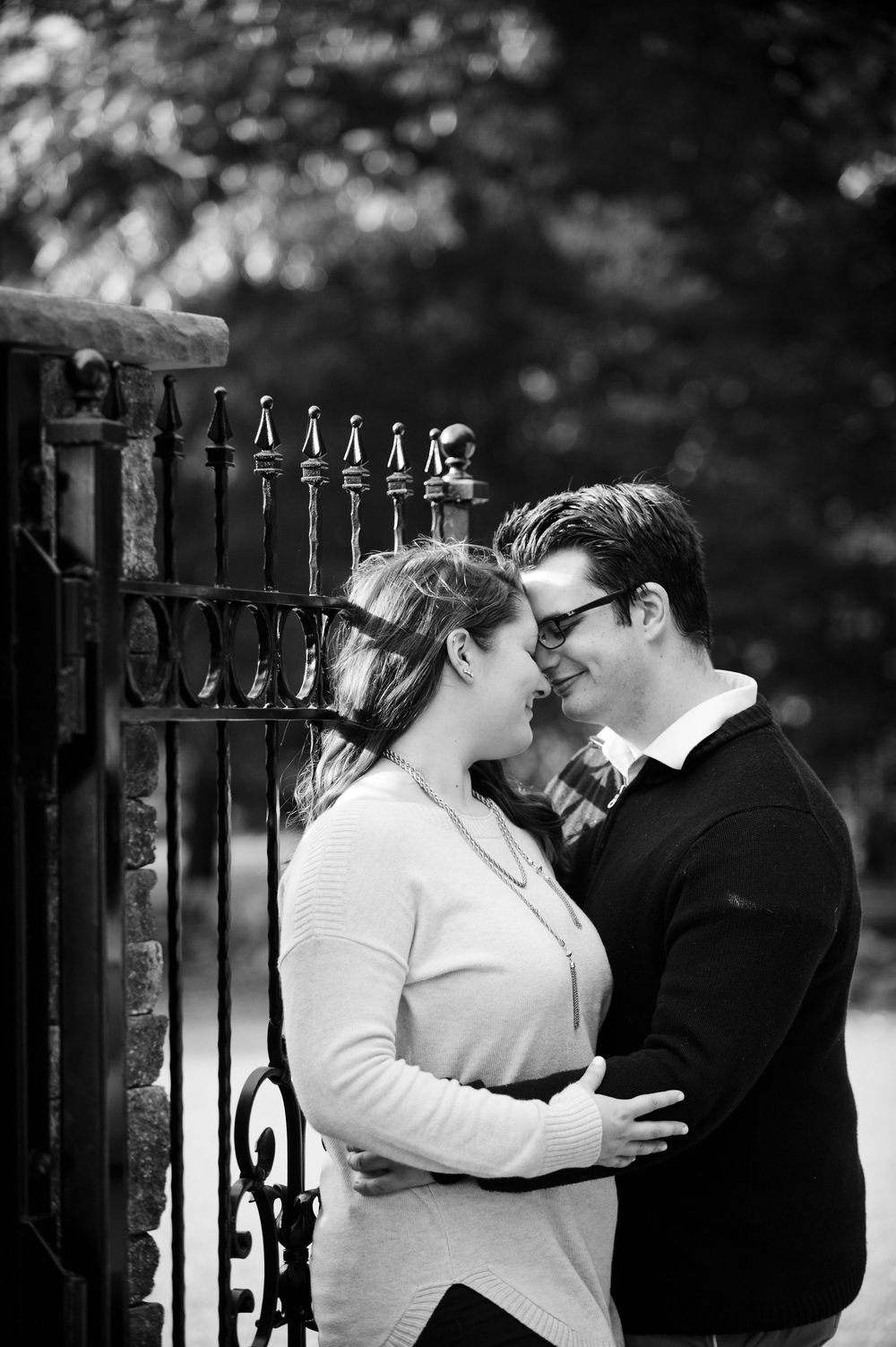 Black and white romantic photo of couple nearly kissing beside ornate iron garden gate.
