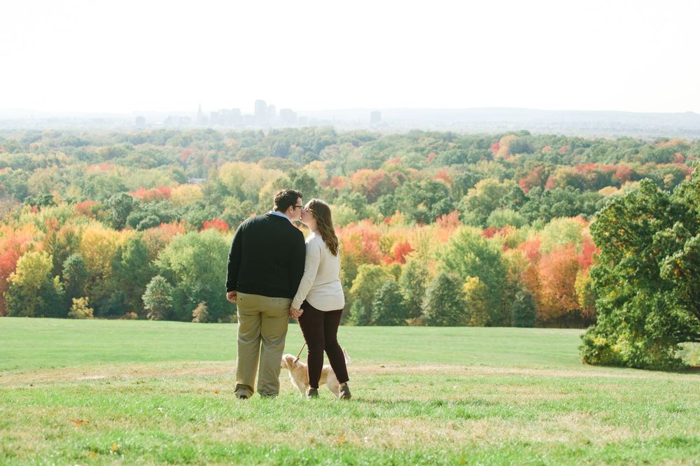 Couple walking with their small dog across a green hill overlooking vibrant autumn foliage.