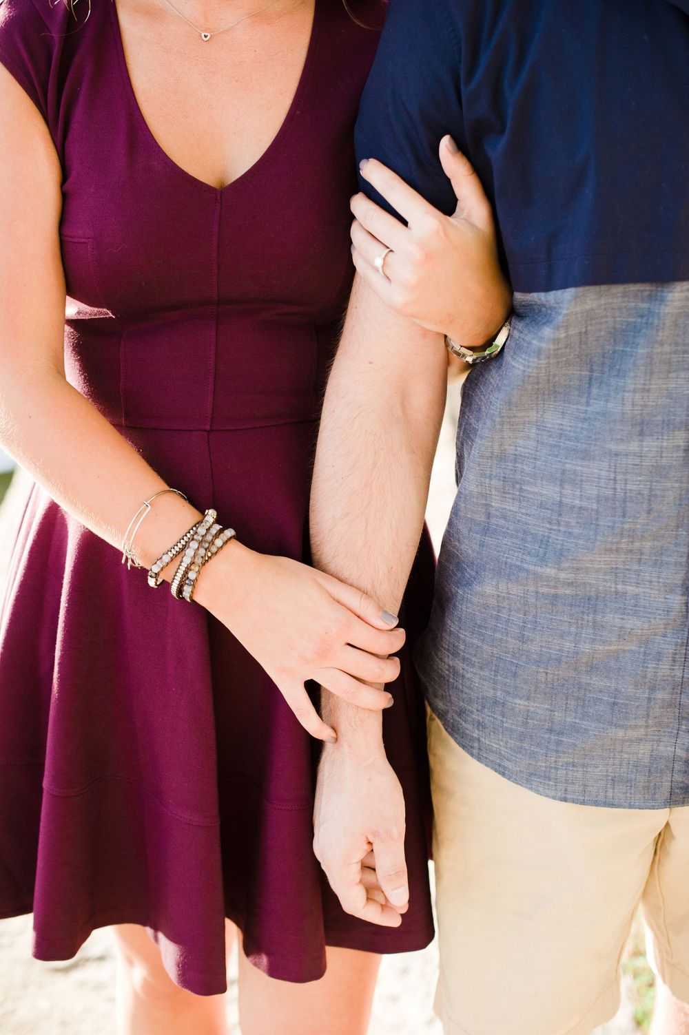 Close-up of a couple holding arms showing an engagement ring, bracelets, and a burgundy dress.