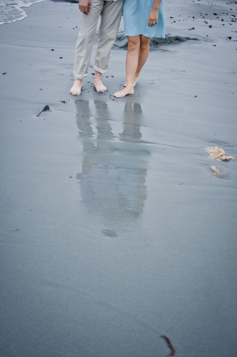 Barefoot couple standing together on wet reflective sand at the beach, wearing casual summer attire.