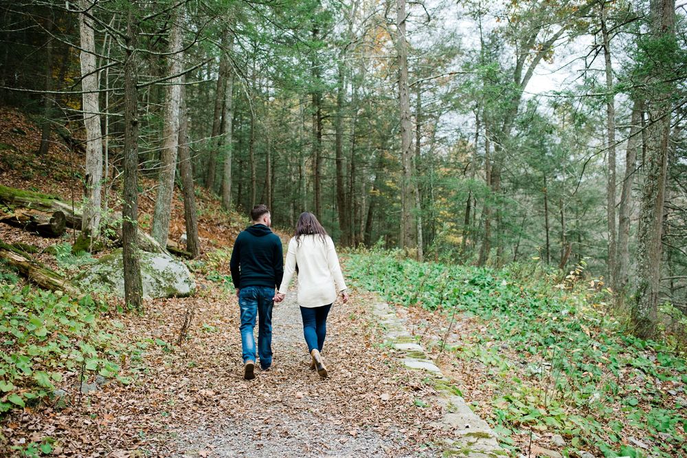 Couple walking hand in hand along a leaf-covered forest path surrounded by tall trees in fall.