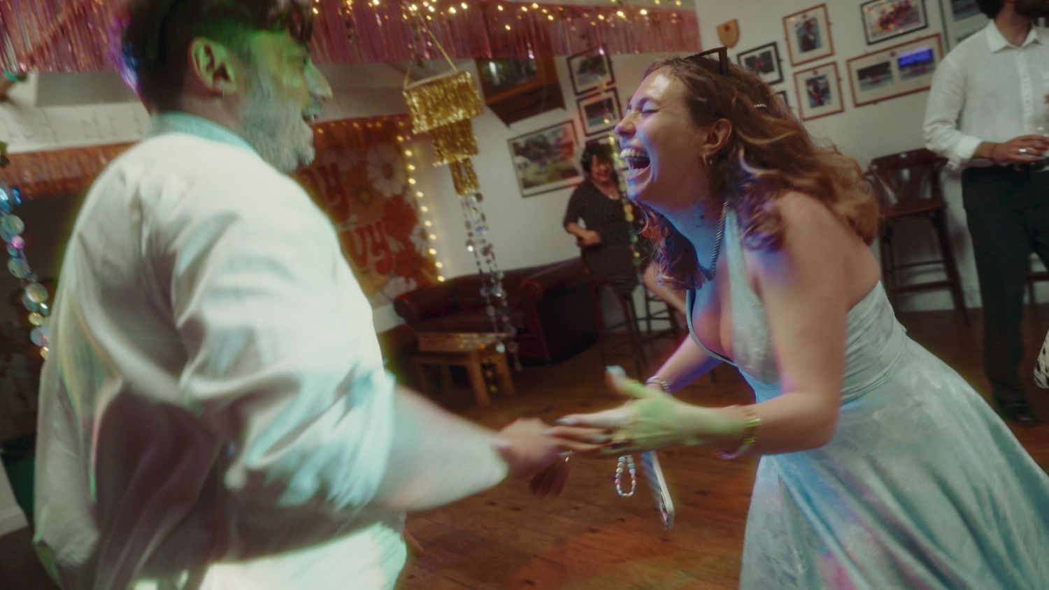 Two people dancing joyfully at a festive indoor party, laughing and holding hands in colorful attire.