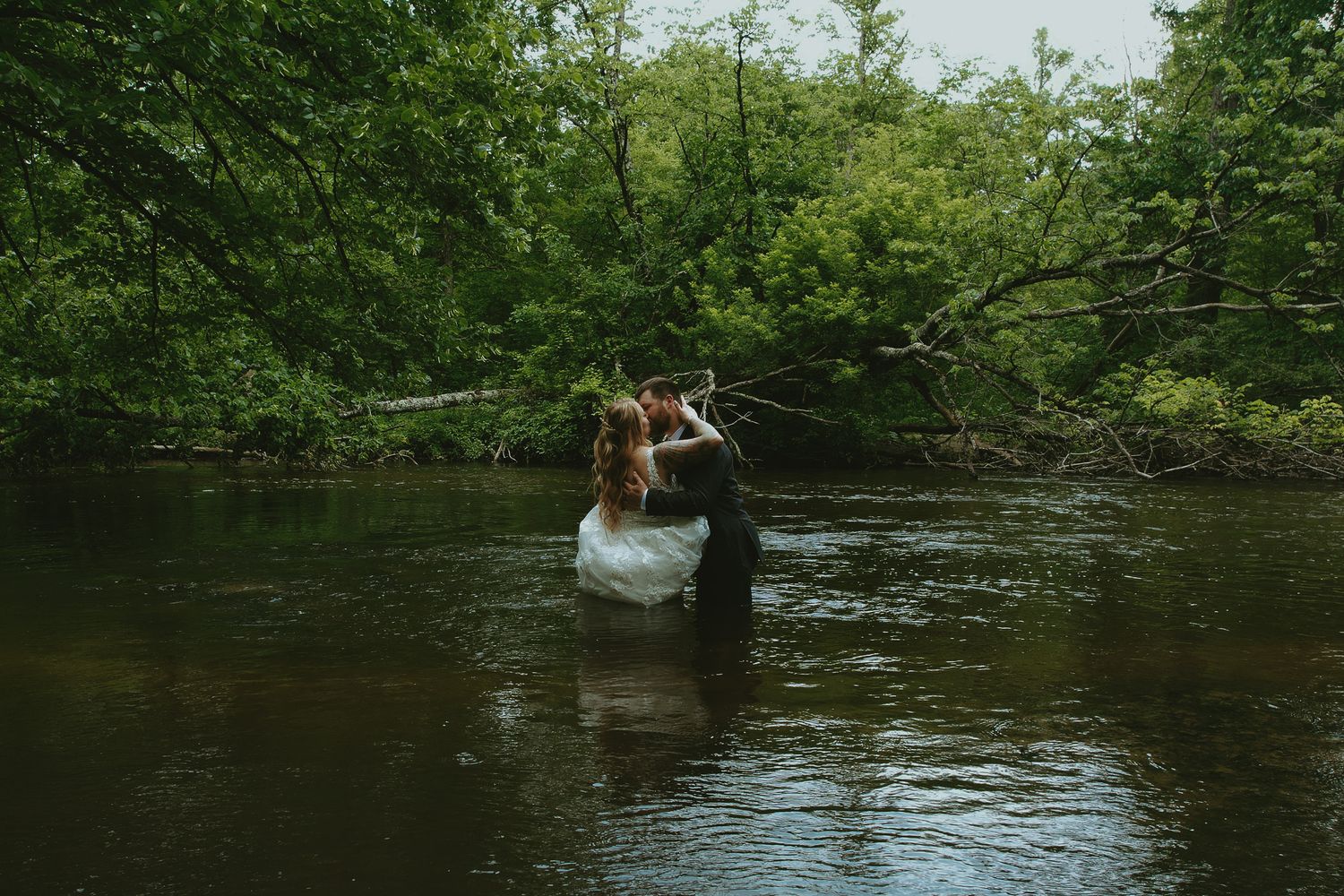Bride and groom sharing a romantic kiss while standing waist-deep in a serene river surrounded by lush green trees.