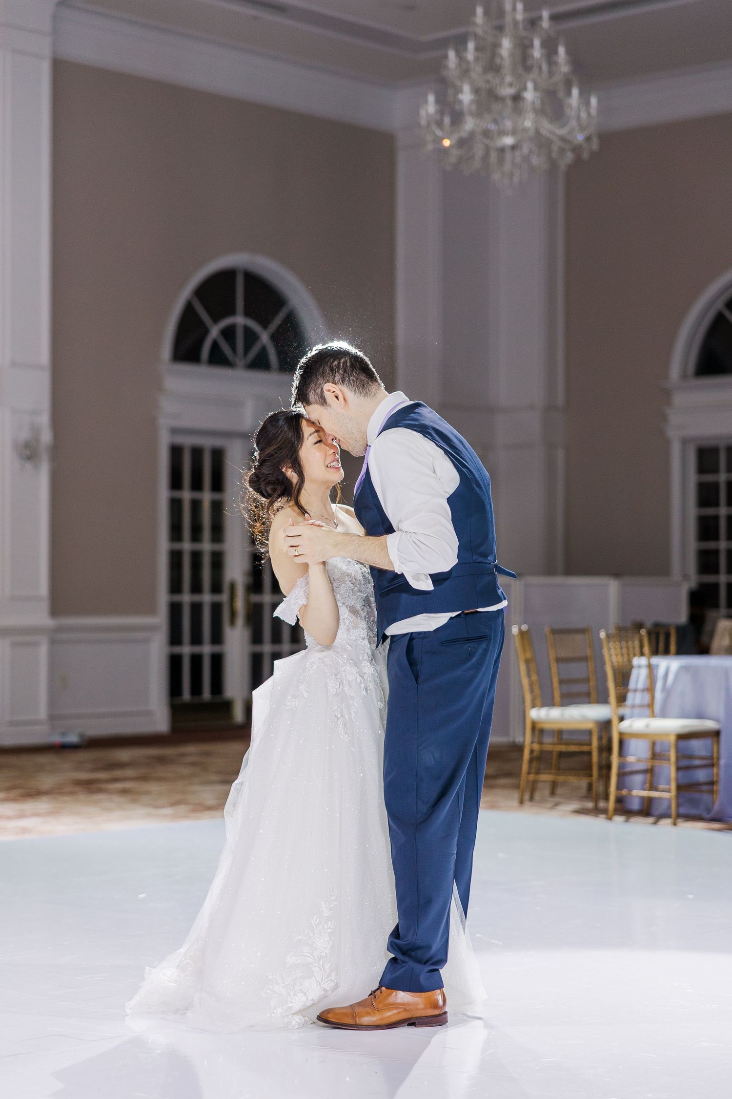 Bride and groom share their first dance in an elegant ballroom with chandelier lighting and white dance floor.