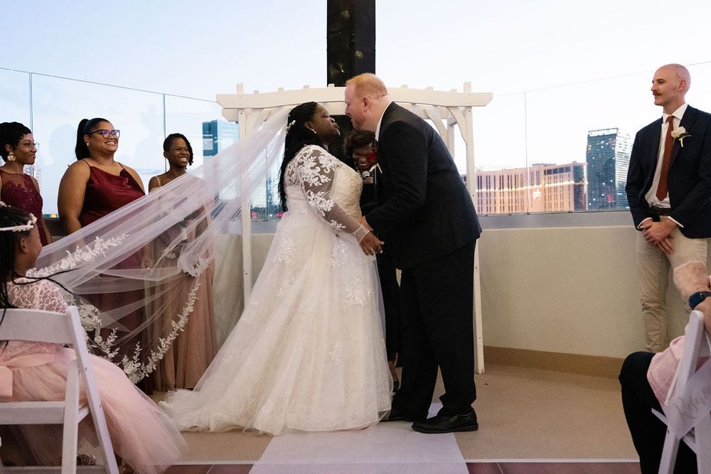 Bride and groom share first kiss at outdoor rooftop wedding ceremony with bridal party in burgundy dresses.