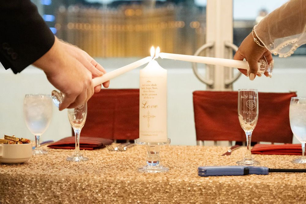Two hands lighting a unity candle at a wedding reception table with champagne flutes and red accents.