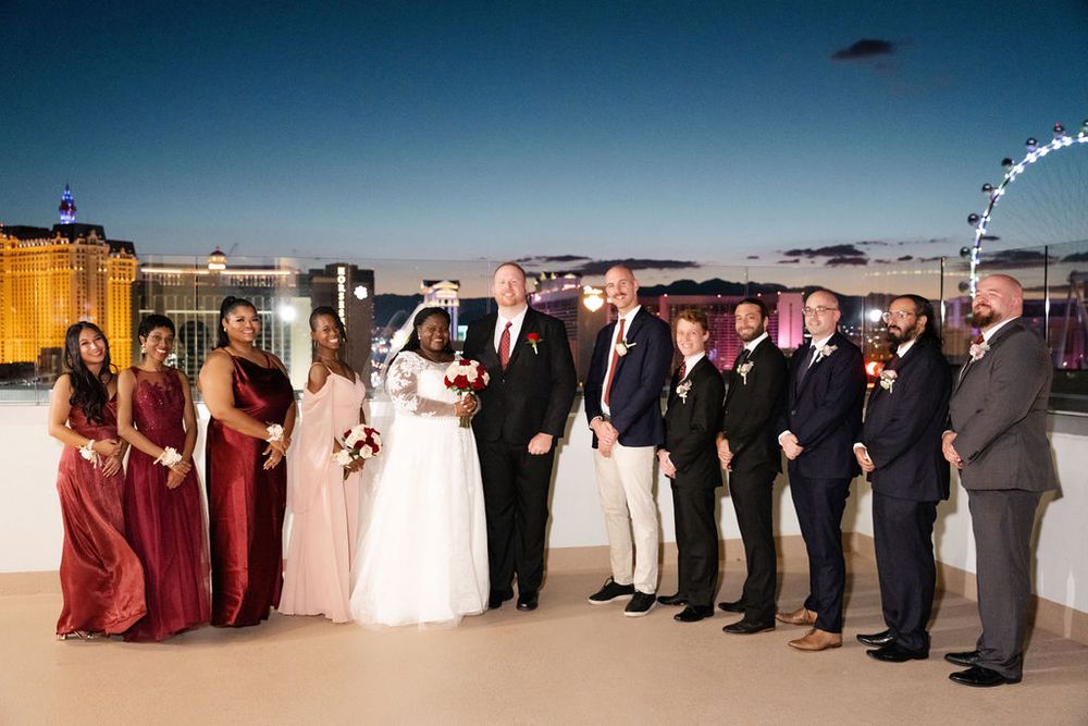 Wedding party poses on rooftop in Las Vegas at dusk, bridesmaids in burgundy gowns and groomsmen in navy suits.