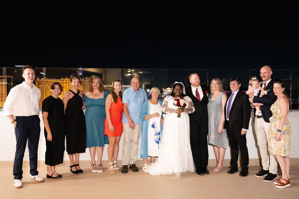 Wedding party poses outdoors at night, bride in white gown with diverse group of guests smiling together.